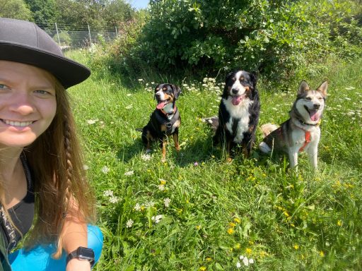 Frau mit langer Haaren posiert lächelnd neben drei Hunden im Gras.